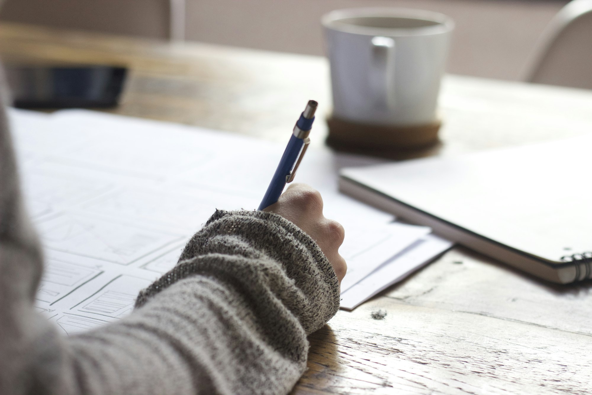 A person writing with a pen in a notebook beside a cup of coffee.